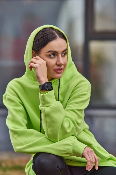Brunette jeune femme en couleur citron vert sweat à capuche posant avec montre intelligente au poignet et regardant loin à l'extérieur — Photo de stock