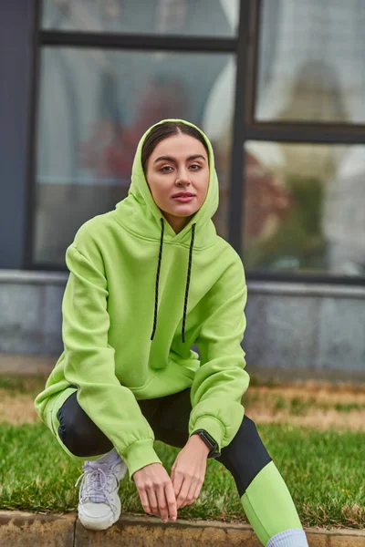 Brunette jeune femme en couleur citron vert sweat à capuche posant avec montre intelligente au poignet et regardant la caméra — Photo de stock