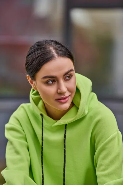 Portrait de jeune femme rêveuse aux cheveux bruns en sweat à capuche de couleur lime regardant vers l'extérieur — Photo de stock