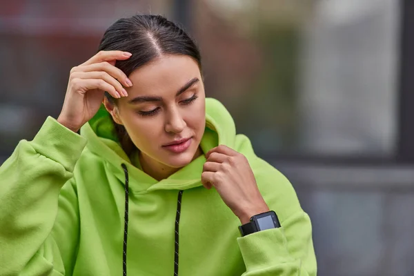 Jolie jeune femme en couleur lime sweat à capuche posant avec montre intelligente au poignet et détournant les yeux — Photo de stock