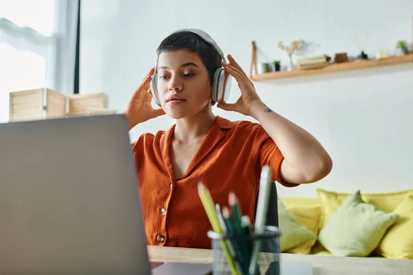 Bonita mujer joven en camisa naranja con auriculares que estudian en su computadora portátil en casa, educación - foto de stock