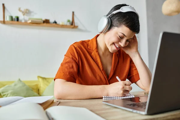 Joyous female student in orange casual shirt with headphones taking notes while studying, education — Stock Photo
