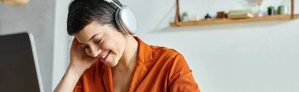 Alegre estudiante de camisa naranja con auriculares estudiando en su portátil, educación, pancarta - foto de stock