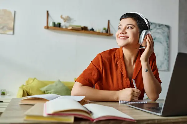 Alegre joven estudiante en naranja camisa escuchar música mientras estudia en el ordenador portátil, mirando hacia otro lado - foto de stock