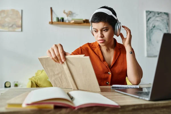 Jeune femme attrayante avec les cheveux courts et écouteurs livre de lecture tout en étudiant à la maison, éducation — Photo de stock