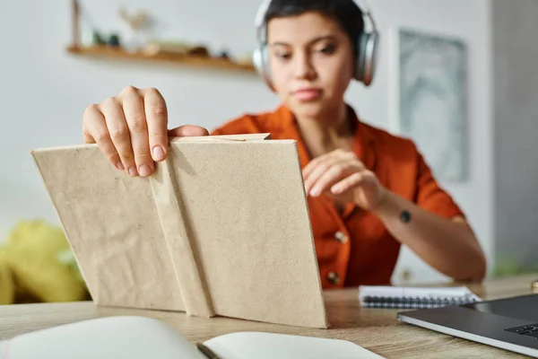 Focus on textbook in hands of blurred young female student with headphones and tattoo, studying — Stock Photo
