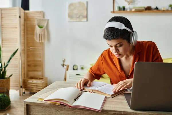 Mujer de pelo corto concentrado con auriculares que estudian desde casa mirando libro de texto, educación - foto de stock