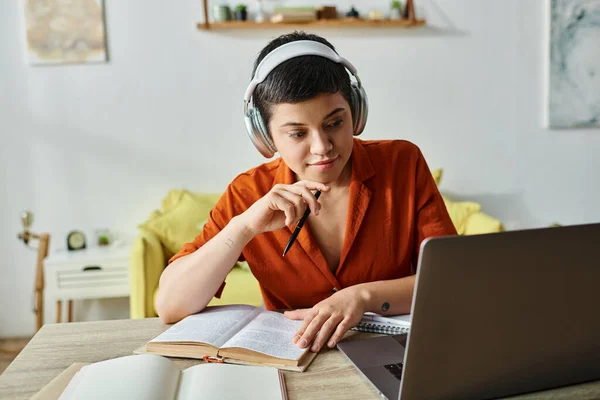 Mujer pensativa en camisa naranja con auriculares y libro de estudio en su ordenador portátil en casa, la educación - foto de stock