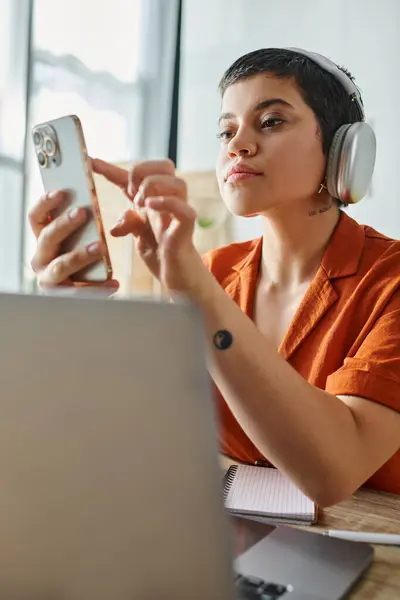 Vertical shot of attractive short haired student with headphones looking at phone in front of laptop — Stock Photo