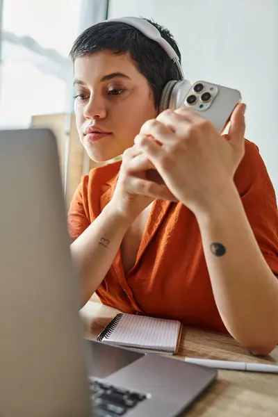 Disparo vertical de mujer joven con teléfono y auriculares mirando cuidadosamente a su computadora portátil, estudiando - foto de stock