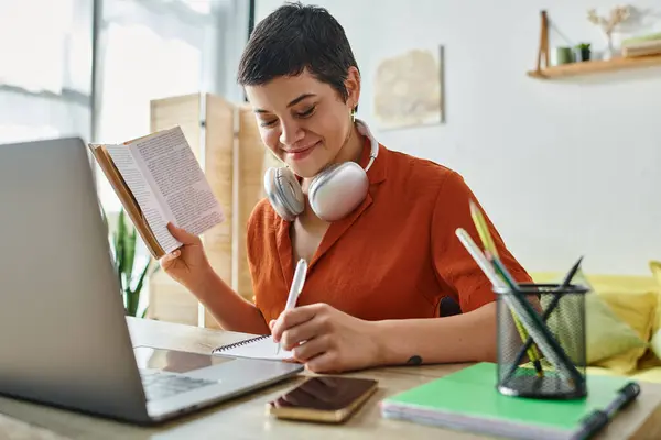 Alegre estudiante de pelo corto con auriculares y libros anotando notas, portátil en la mesa, estudiando - foto de stock