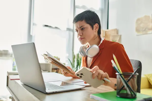 Mujer enfocada con pelo corto y tatuajes sosteniendo el teléfono móvil y mirando a la computadora portátil, estudiando - foto de stock