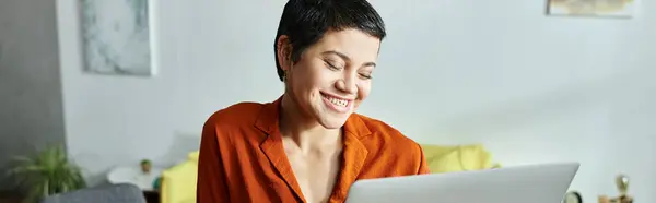 Feliz estudiante atractiva con piercing en camisa naranja sonriendo a su computadora portátil, educación, pancarta - foto de stock