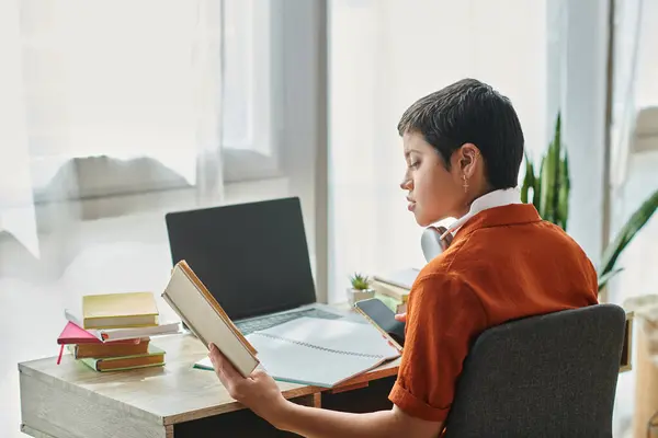 Estudiante enfocado con libro de texto de lectura de pelo corto con teléfono en las manos y auriculares, educación - foto de stock