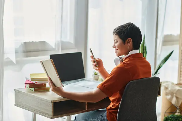 Alegre joven estudiante mirando el libro de texto con el teléfono en las manos y portátil en la mesa, estudiando - foto de stock