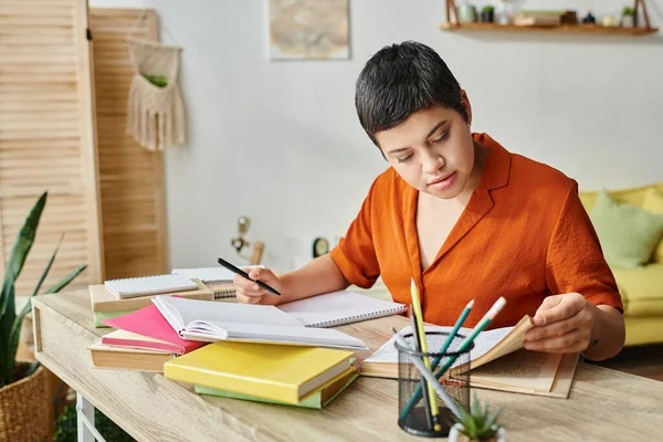 Estudiante joven trabajador en camisa naranja tomando notas y mirando el libro de texto, la educación en casa - foto de stock