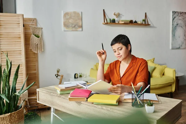 Fröhliche junge Frau in lässiger Hauskleidung, die hart am Schreibtisch lernt und sich Bücher ansieht, Bildung zu Hause — Stockfoto