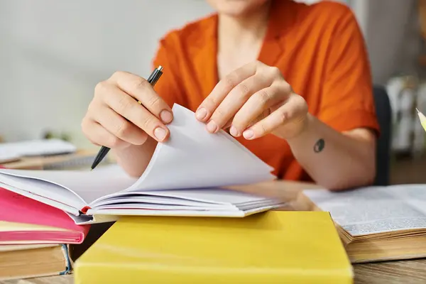 Recortado vista de joven estudiante con tatuaje en naranja camisa dando vuelta páginas de libro de texto, estudiando desde casa - foto de stock