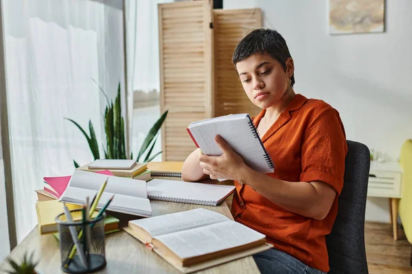 Atractiva joven estudiante mirando atentamente sus notas sentado en el escritorio, la educación en casa - foto de stock