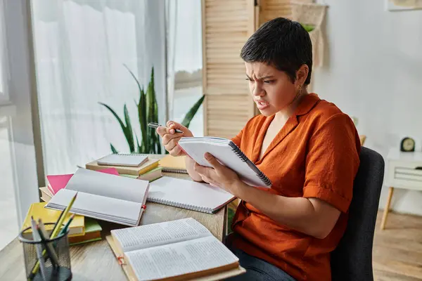 Confundida estudiante de pelo corto en camisa naranja mirando su libro de texto mientras estudia duro - foto de stock