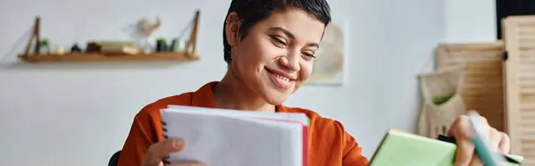 Alegre joven estudiante sentada en la mesa y mirando sus notas y libros, educación, pancarta - foto de stock