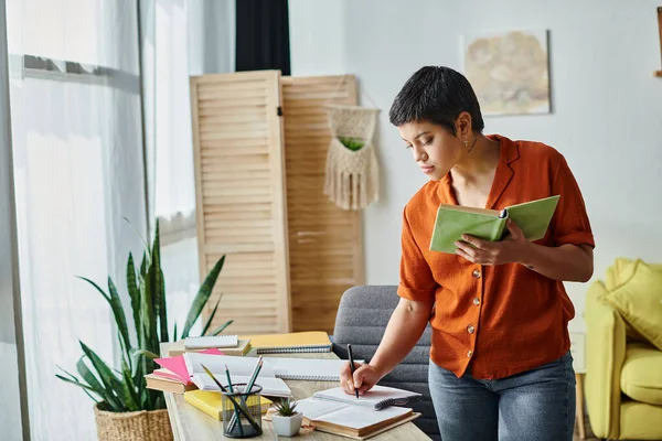 Estudiante atractivo enfocado en camisa naranja de pie cerca de escritorio sosteniendo libro tomando notas, educación - foto de stock