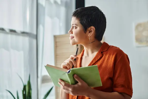 Atractiva mujer alegre sosteniendo libro de texto y pluma y mirando soñadoramente lejos, la educación en el hogar - foto de stock