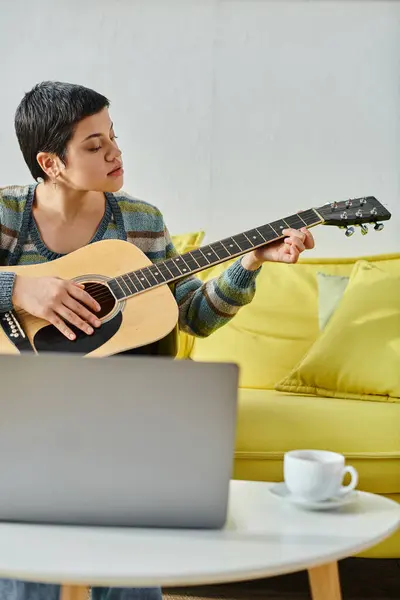 Vertikale Aufnahme einer jungen kurzhaarigen Frau, die vor dem Laptop sitzt und lernt, wie man Gitarre spielt — Stockfoto
