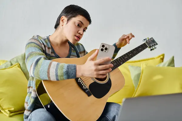 Mujer enfocada de buen aspecto que asiste a clase de guitarra en línea con teléfono en las manos, educación en casa - foto de stock