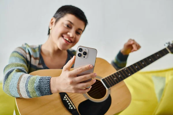 Mujer atractiva joven mirando alegremente al teléfono móvil con la guitarra en las manos, la educación en el hogar - foto de stock