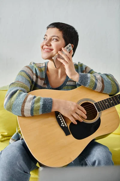 Tiro vertical de mujer alegre con piercing sosteniendo la guitarra y hablando por teléfono, educación en casa - foto de stock