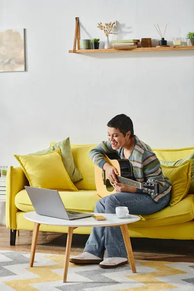 Vertical shot of joyful young woman sitting on sofa with guitar looking on laptop on remote class — Stock Photo