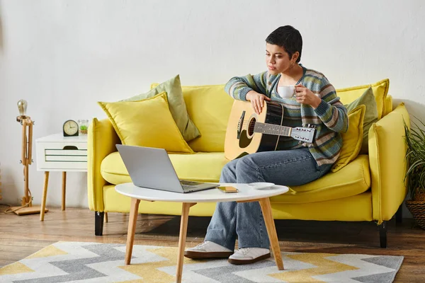 Концентрированная молодая женщина, сидящая на диване с гитарой во время урока онлайн музыки, образование дома — стоковое фото