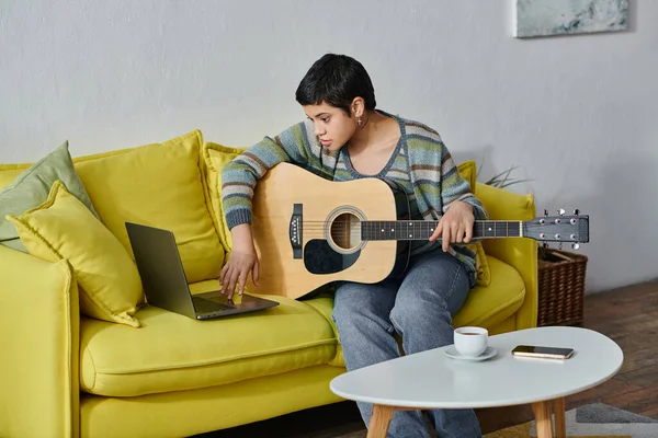 Atractiva joven sentada en el sofá con la guitarra en las manos y mirando a la computadora portátil, la educación en el hogar - foto de stock