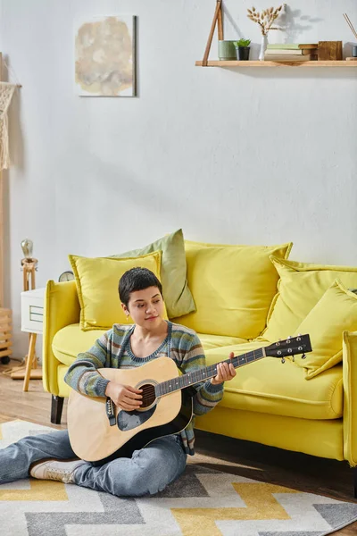 Tiro vertical de una joven guapa tocando la guitarra y sentada en el suelo, educación en casa - foto de stock