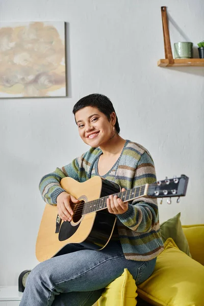 Tiro vertical de sonriente mujer feliz en traje casual tocando la guitarra en el sofá, la educación en casa - foto de stock