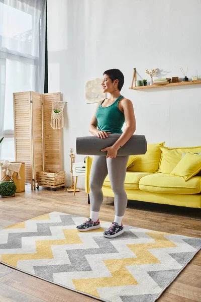 Bella donna atletica con tappetino fitness in piedi di fronte al divano giallo, fitness e sport — Foto stock