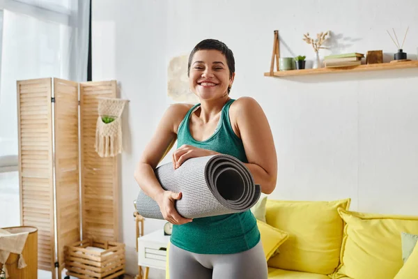 Mujer atractiva feliz en ropa deportiva sonriendo alegremente a la cámara con alfombra de fitness en las manos - foto de stock