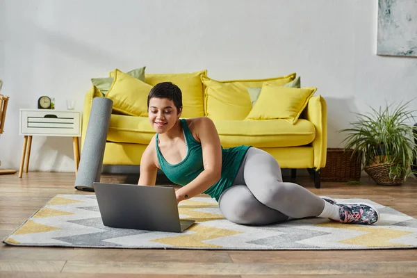 Mujer alegre con el pelo corto acostado en el suelo y mirando a la computadora portátil durante la lección de deporte remoto - foto de stock