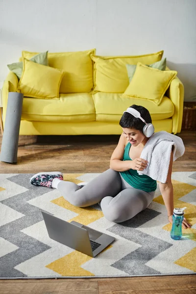 Plan vertical d'une femme joyeuse assise sur le sol avec une serviette à côté d'un ordinateur portable et d'une bouteille d'eau, fitness — Photo de stock