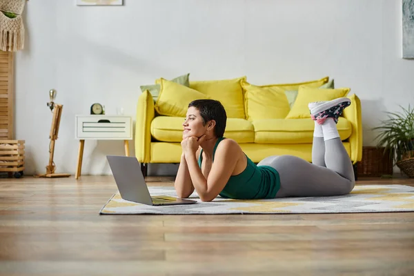 Jolly donna sdraiata sul pavimento durante la lezione di fitness online sorridente felicemente, mani sotto il mento, fitness — Foto stock