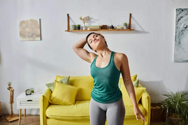 Mujer joven de buen aspecto con el pelo corto que estira su cuerpo delante del sofá amarillo, fitness - foto de stock