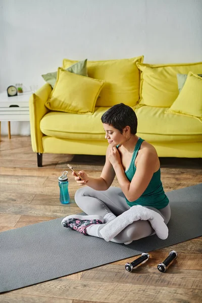 Colpo verticale di bella donna bruna seduta sul pavimento e guardando il telefono cellulare, fitness — Foto stock