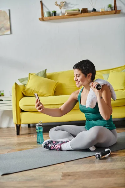 Plan vertical de jeune femme heureuse assise sur le sol avec haltère et serviette prendre selfie, fitness — Photo de stock