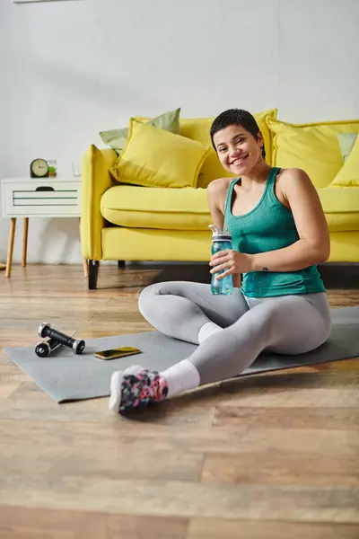 Vertical shot of cheerful woman sitting on floor with water bottle in hands and smiling at camera — Stock Photo