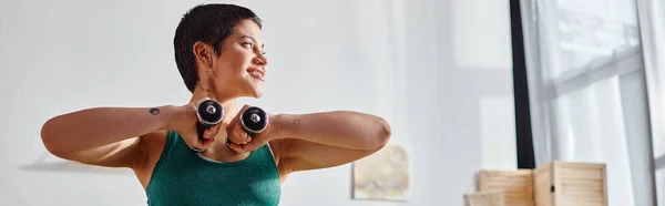 Mujer alegre con pelo corto haciendo ejercicio con pesas y mirando hacia otro lado, fitness y deporte, pancarta - foto de stock