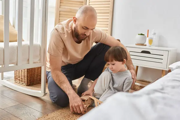 Красивий бородатий батько грає з дерев'яними іграшками зі своїм маленьким сином поруч з ліжечком, сімейна концепція — стокове фото