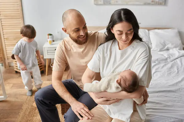 Focus on modern loving parents holding their newborn baby with their blurred son playing on backdrop — Stock Photo