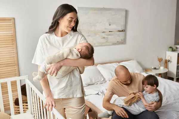 Glückliche Mutter lächelt ihr Neugeborenes an, das neben der Krippe steht und ihr Mann mit Sohn spielt — Stockfoto