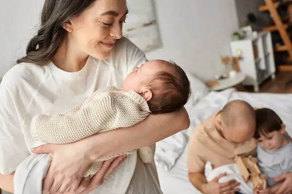 Зосередьтеся на щасливій матері, дивлячись на її новонародженого з розмитим чоловіком і маленьким сином, який грає на тлі — стокове фото
